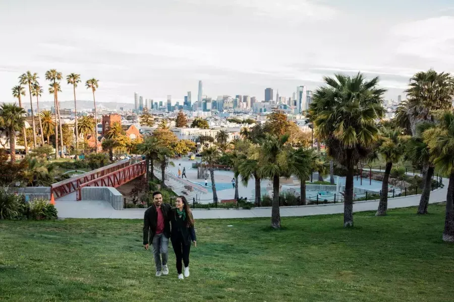 Un couple se dirige vers la caméra avec Dolores Park et la Skyline de San Francisco derrière eux.